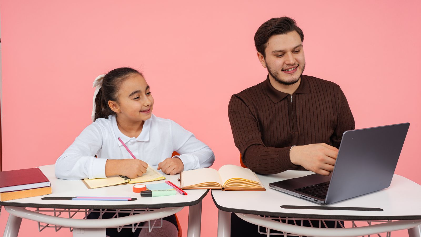 A teacher guides a student in a classroom with books and a laptop on a pink background.
